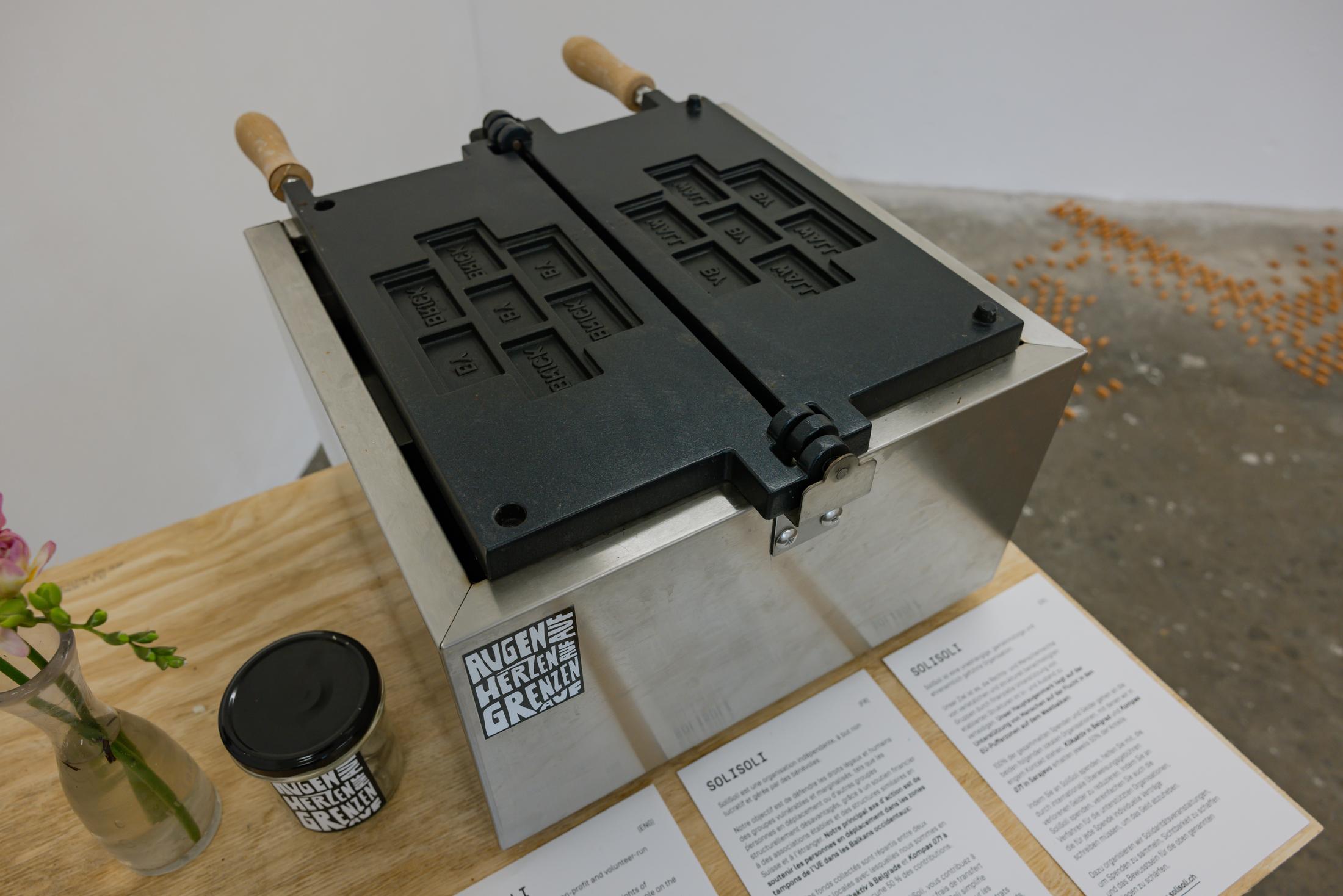 Exhibition View Duo Show Stirnimann-Stojanovic «Connecting through Collective Care and Redistribution; view on Stirnimann-Stojanovic X SoliSoli, SoliSoliWaffle, Custom-made waffle machine (the waffles mimic a brick wall inscribed with “Brick by Brick” and “Wall by Wall”), 37.5 x 50 x 27 cm, 2024» at WallStreet Fribourg, Fribourg, 2025 / Photo: Julie Folly / Courtesy: the artists and WallStreet